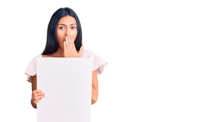 Young beautiful latin girl holding blank empty banner covering mouth with hand, shocked and afraid for mistake. surprised expression