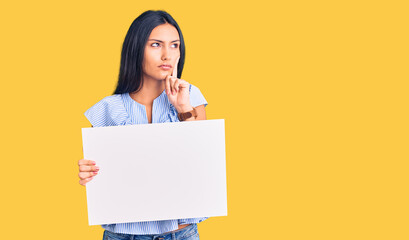 Young beautiful latin girl holding blank empty banner serious face thinking about question with...