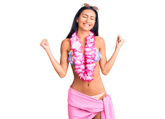 Obraz na płótnie Canvas Young beautiful latin girl wearing bikini and hawaiian lei very happy and excited doing winner gesture with arms raised, smiling and screaming for success. celebration concept.