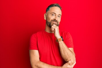 Handsome middle age man wearing casual red tshirt smiling looking confident at the camera with crossed arms and hand on chin. thinking positive.