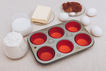 Red paper molds, metal baking tray, eggs, glass of flour, glass of milk, cacao and piece of butter on tablecloth. Ingredients for cupcakes. 