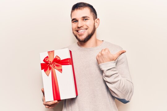 Young handsome man holding gift pointing thumb up to the side smiling happy with open mouth