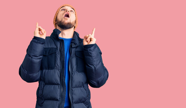 Young handsome man wearing winter clothes amazed and surprised looking up and pointing with fingers and raised arms.