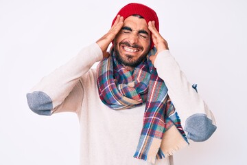 Handsome young man with curly hair and bear wearing a winter sweater, scarf and wool hat with hand on head, headache because stress. suffering migraine.