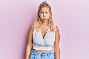 Young blonde girl wearing casual clothes puffing cheeks with funny face. mouth inflated with air,...