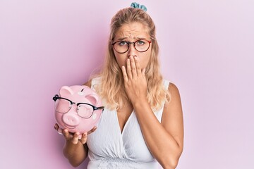 Young blonde girl holding piggy bank with glasses covering mouth with hand, shocked and afraid for...