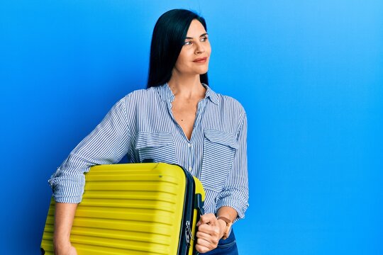 Young caucasian woman holding cabin bag smiling looking to the side and staring away thinking.