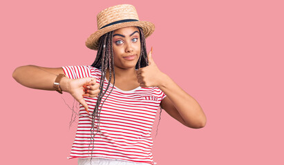 Young african american woman with braids wearing summer hat doing thumbs up and down, disagreement and agreement expression. crazy conflict