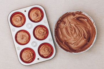 Cacao dough for cupcakes in bowl and red paper molds with dough in metal baking tray on tablecloth. Preparing process to baking. Top view.