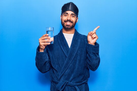 Young hispanic man wearing sleep mask and robe drinking water smiling happy pointing with hand and finger to the side