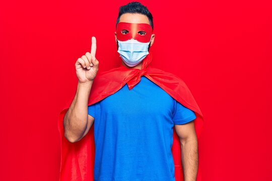 Young hispanic man wearing super hero costume and medical mask smiling with an idea or question pointing finger with happy face, number one