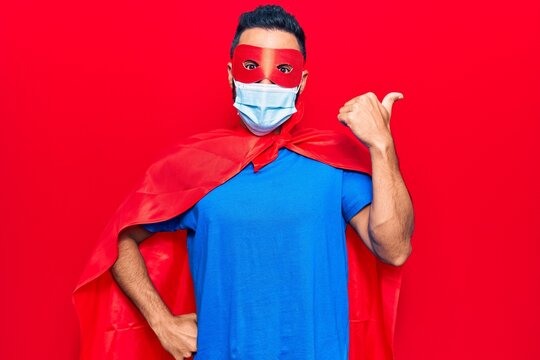 Young hispanic man wearing super hero costume and medical mask pointing thumb up to the side smiling happy with open mouth