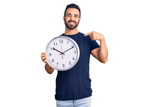 Young hispanic man holding big clock pointing finger to one self smiling happy and proud