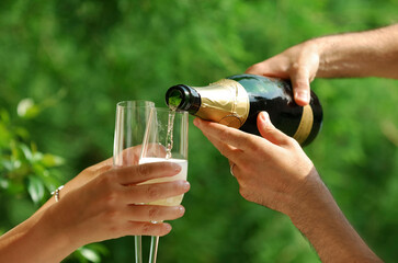 Hands of man and woman preparing to celebrate with champagne on out of focus green lawn background. Man hands pouring champagne in crystal glasses hold by female or woman hands at wedding party