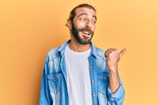 Attractive man with long hair and beard wearing casual denim jacket pointing thumb up to the side smiling happy with open mouth