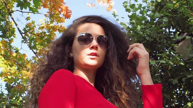 Attractive young brunette woman in sunglasses standing in the forest and touching her hair looks at the camera. Woman is smiling to the camera. Slow motion