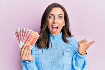 Young brunette woman holding norwegian krone banknotes pointing thumb up to the side smiling happy with open mouth