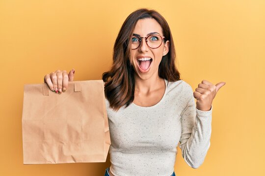 Young brunette woman holding take away paper bag pointing thumb up to the side smiling happy with open mouth