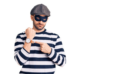 Young handsome man wearing burglar mask in hurry pointing to watch time, impatience, looking at the...