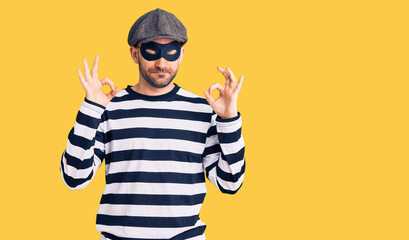 Young handsome man wearing burglar mask relaxed and smiling with eyes closed doing meditation gesture with fingers. yoga concept.