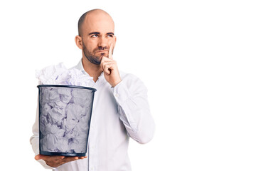 Young handsome man holding paper bin full of crumpled papers serious face thinking about question...