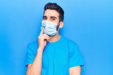 Young handsome man with beard wearing medical mask smiling looking confident at the camera with crossed arms and hand on chin. thinking positive.