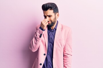 Young handsome man with beard wearing elegant jacket smelling something stinky and disgusting,...