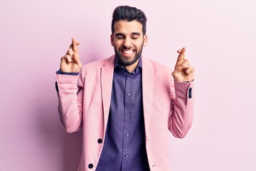 Young handsome man with beard wearing elegant jacket gesturing finger crossed smiling with hope and...