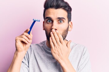 Young handsome man with beard holding razor covering mouth with hand, shocked and afraid for mistake. surprised expression