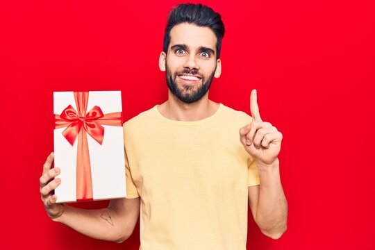 Young handsome man with beard holding gift smiling with an idea or question pointing finger with happy face, number one
