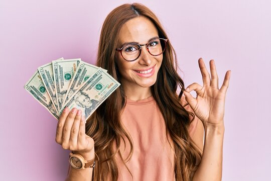 Young brunette woman holding dollars doing ok sign with fingers, smiling friendly gesturing excellent symbol