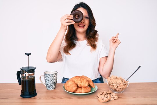 Young beautiful brunette woman eating breakfast holding cholate donut smiling happy pointing with hand and finger to the side