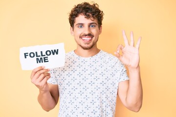 Young caucasian man with curly hair holding follow me message paper doing ok sign with fingers, smiling friendly gesturing excellent symbol