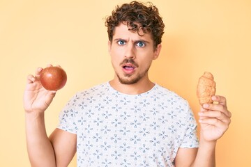 Young caucasian man with curly hair holding apple and croissant in shock face, looking skeptical and sarcastic, surprised with open mouth