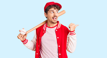 Young handsome man with curly hair playing baseball holding bat and ball pointing thumb up to the side smiling happy with open mouth