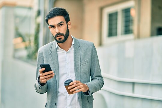 Young hispanic businessman with serious expression using smartphone and drinking coffee at the city.