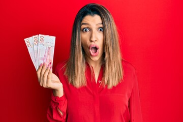 Beautiful brunette woman holding 500 swedish krona banknotes scared and amazed with open mouth for surprise, disbelief face