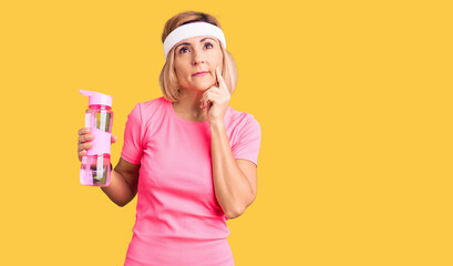Young blonde woman wearing sportswear holding water bottle serious face thinking about question...