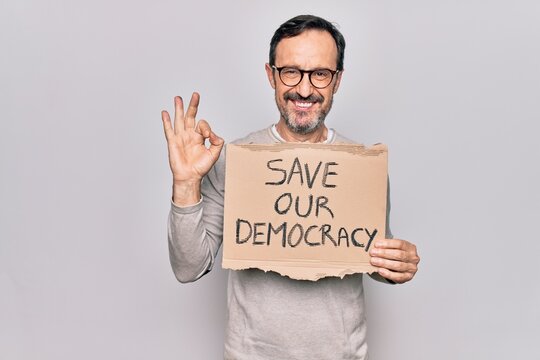 Middle age man holding banner with saver our democracy message over white background doing ok sign with fingers, smiling friendly gesturing excellent symbol