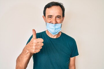 Middle age hispanic man wearing medical mask smiling happy and positive, thumb up doing excellent and approval sign
