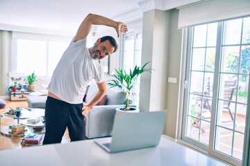 Middle age man with beard training and stretching doing exercise at home looking at sport video on computer