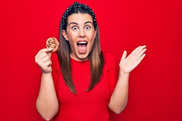 Young beautiful brunette woman holding sweet chocolate cookie over isolated red background celebrating achievement with happy smile and winner expression with raised hand