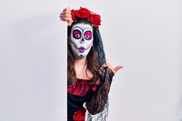 Young woman wearing day of the dead custome holding blank empty banner surprised pointing with hand finger to the side, open mouth amazed expression.