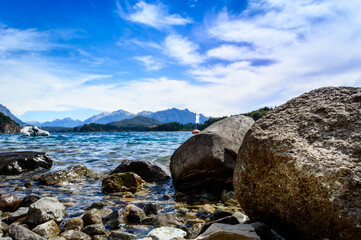 Beautiful landscape seen from the ground behind a large rock. Crystal clear water, mountains, pine trees and rocks. Beach in Bariloche, Argentina on a very sunny day.