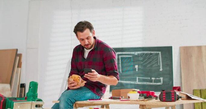 A carpenter uses a smartphone during a break at work. Young handsome craftsman is resting in a carpentry workshop. There are a locksmith table and tools in the background.