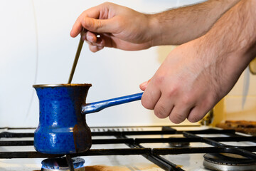 Men's hands prepare coffee in a blue enameled Turk on the gas, stirring with a long teaspoon