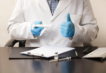 Doctor with blue gloves and airplane in one hand, in the other doing ok, desk with syringe, glasses, white form and computer keyboard