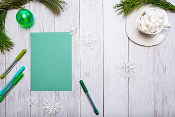 Blank green sheet of paper, green markers and a Christmas ball with spruce branches, white cup with marshmallows on a white wooden background. Cozy festive concept with place for text