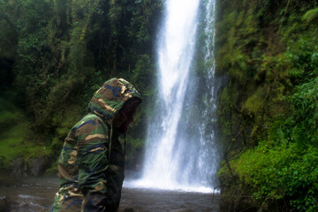 Woman with green camouflage jacket standing in the rocks, in front of a waterfall and rocks with green plants and moss