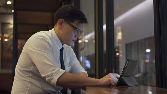 Asian businessman office worker sitting by the window in coffee shop using digital tablet with internet for business online working at coffee shop. Male entrepreneur waiting for partnership in cafe.